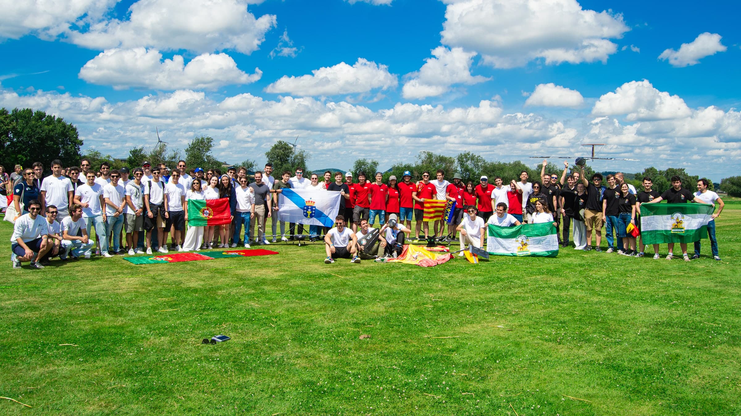 Group photo on an airfield lawn with the Spanish and Portuguese teams at the Air Cargo Challenge 2024, holding the flags of Portugal, Galicia, Spain and Andalusia.